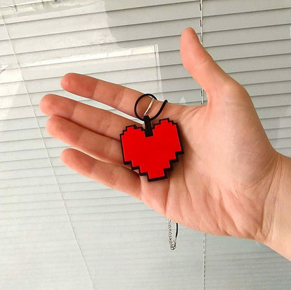 Hand holding a red pixelated heart keychain from Undertale against a window blind background